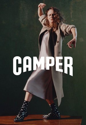 Mujer con abrigo y vestido beige en una pose de danza dinámica, luciendo botas negras con lunares, sobre una plataforma de madera contra un fondo verde oscuro.