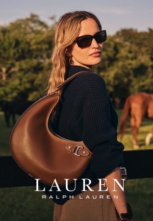 Borsa hobo in pelle marrone con una texture liscia, forma curva e dettaglio con fibbia. Modella che indossa un maglione lavorato a maglia blu navy, all'aperto.