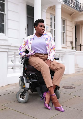 Elektryk rullstol med mörka sittplatser, person klädd i en blommig kavaj, lila topp, bruna byxor och lila klackar, poserad mot arkitektur.