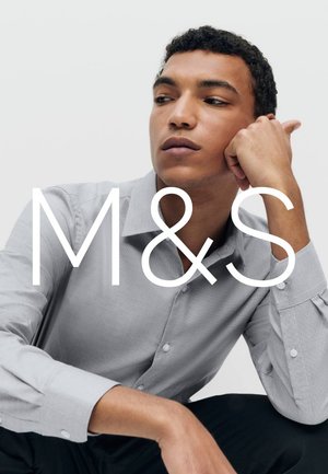 Młody mężczyzna w jasnoszarej koszuli zapinanej na guziki, siedzący z ręką opartą na twarzy, patrzący w bok na jednolitym tle.