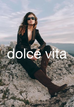 Braune knöchelhohe Stiefel mit einer Wildleder-Textur, getragen zu einem schwarzen maßgeschneiderten Anzug und großen schwarzen Sonnenbrillen, sitzend auf steinigem Gelände unter einem bewölkten Himmel.