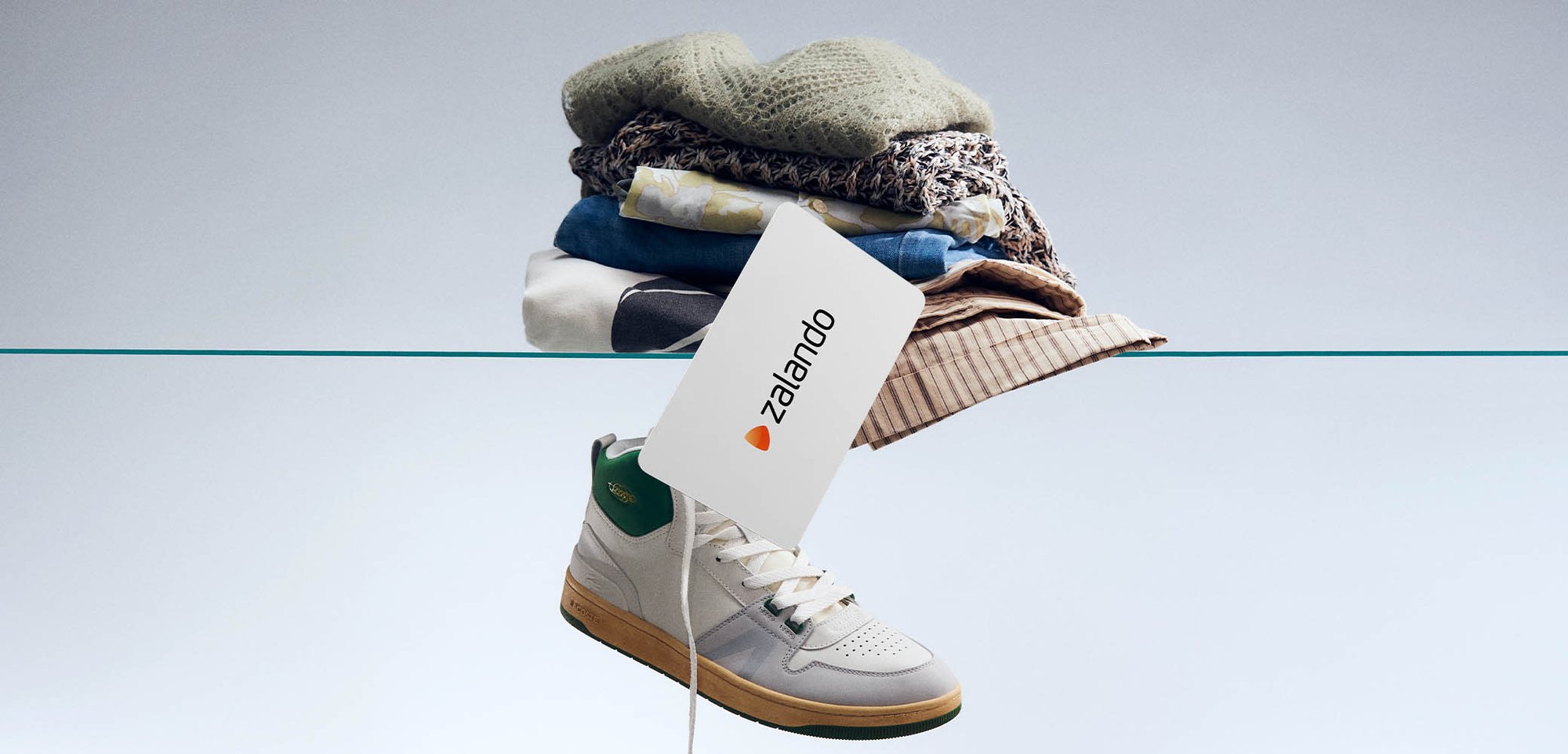 Lacoste high-top sneaker in white and green, featuring a textured upper and flat laces, beside a stack of folded clothing and a Zalando tag.