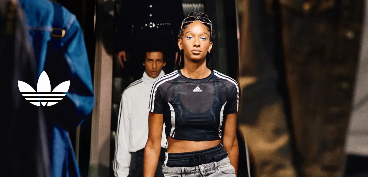 Female model walks runway in black Adidas crop top and gray pants, with male model in white jacket behind her during fashion show.