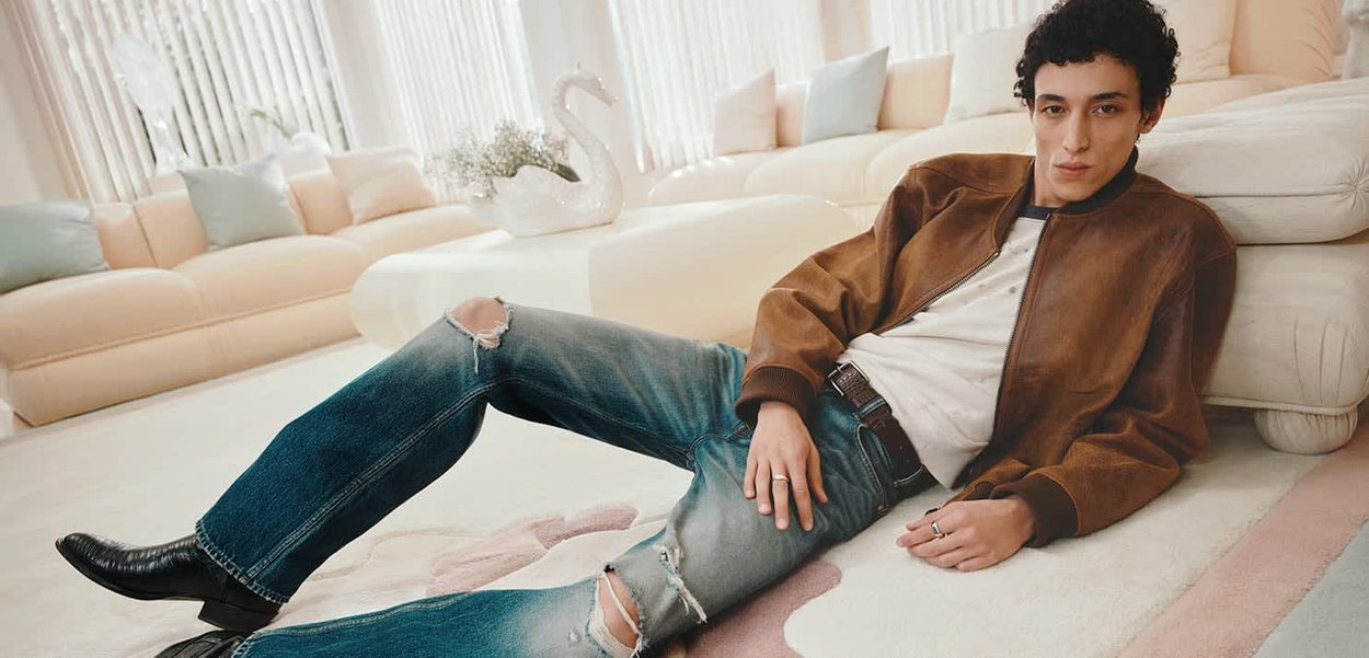 Junger Mann in zerrissenen Jeans und brauner Jacke, der auf dem Teppichboden in einem hellen Wohnzimmer mit hellfarbigen Sofas liegt.