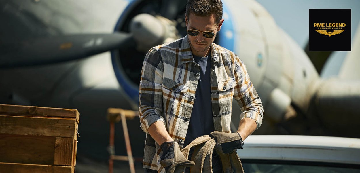 Mann mit Sonnenbrille und kariertem Hemd hält ein Seil vor einem Vintage-Flugzeug, in der Nähe befinden sich Holzkisten und das PME Legend-Logo ist sichtbar.