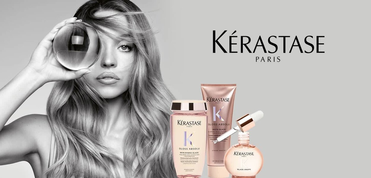 Mujer con cabello largo y ondulado sostiene una esfera de vidrio cerca del ojo, junto a productos para el cuidado del cabello Kérastase París en envases de color rosa suave.