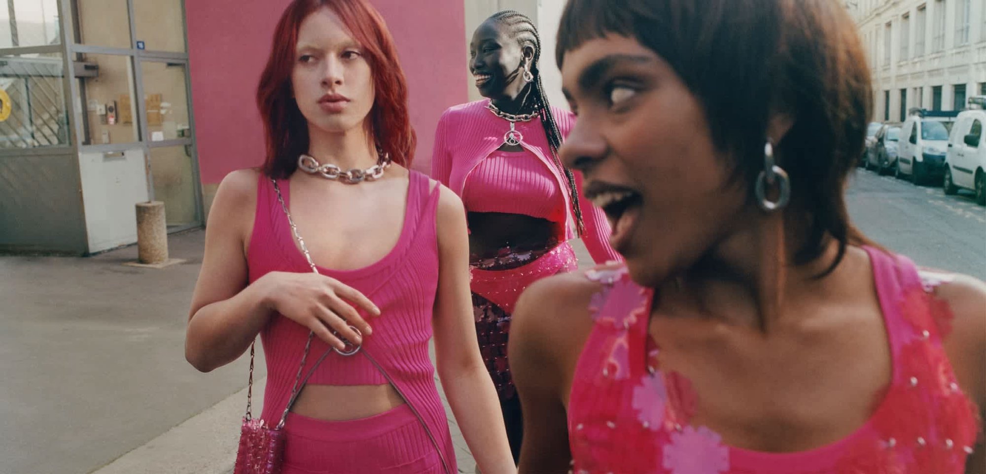 Tre persone indossano abiti di un vivace rosa. Maglie a coste e pantaloni attillati. Gli accessori includono catene e una borsa di perline.