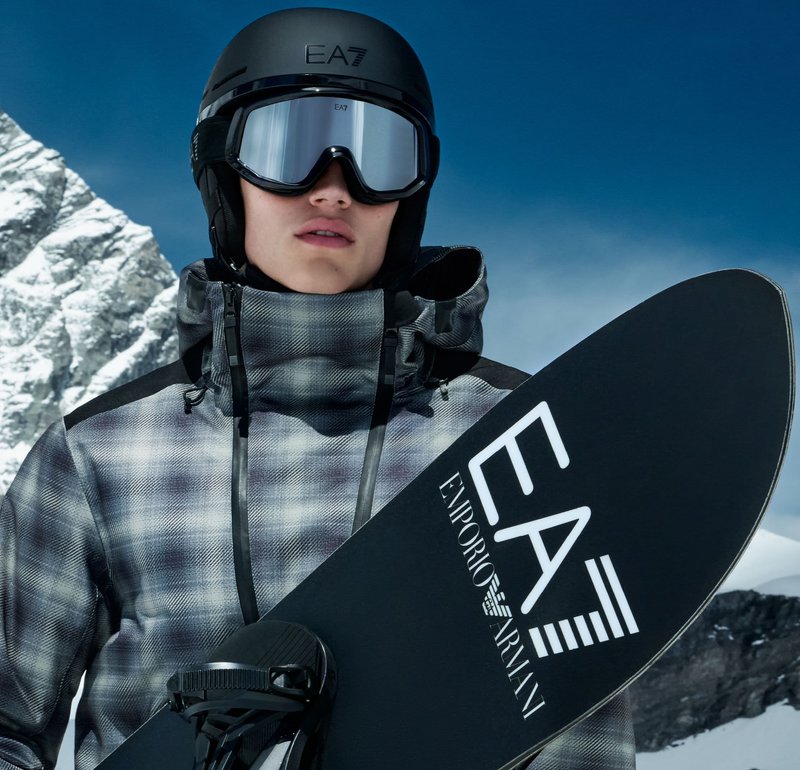 Snowboarder trägt schwarzen Helm, verspiegelte Skibrille und grau gemusterten Anorak, hält ein schwarzes Snowboard mit weißen Logoakzenten.