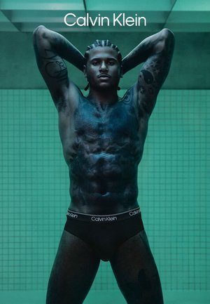 Schwarze Calvin Klein Slips getragen von einer muskulösen Figur mit sichtbaren Tätowierungen, die in einer coolen, gefliesten Umgebung mit gehobenen Armen steht.