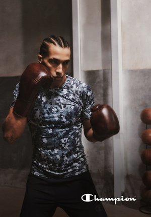 L'abbigliamento sportivo presenta una maglietta aderente a maniche corte con motivo camouflage in tonalità di grigio e blu, abbinata a guantoni da boxe marroni.