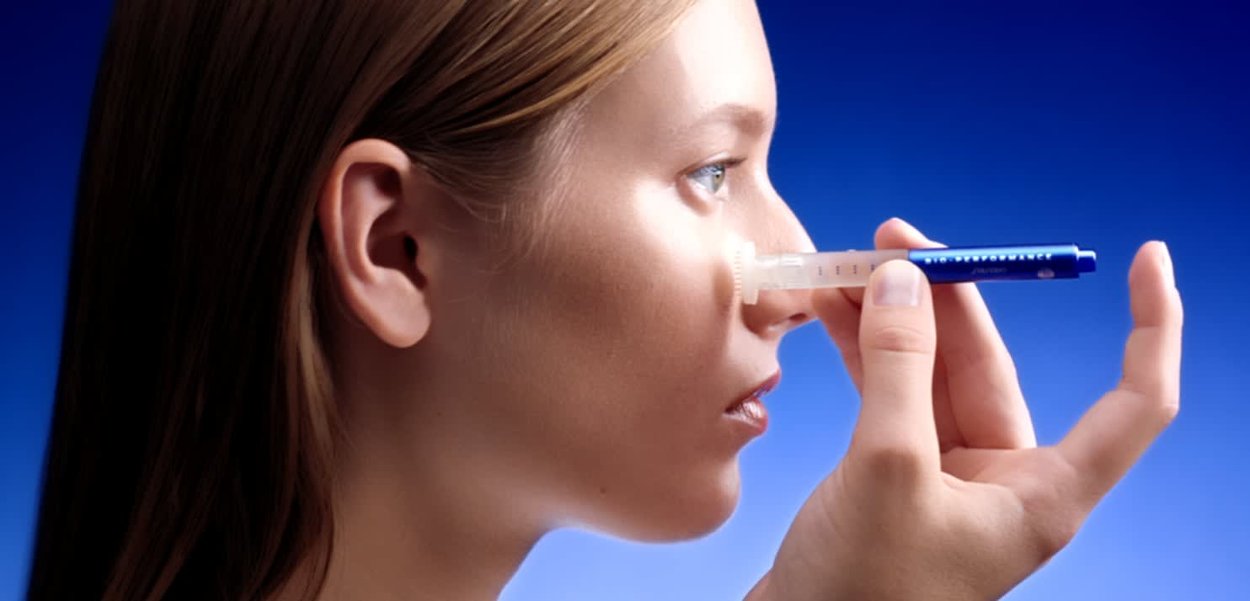 Een heldere neussprayhouder die dichtbij de neus wordt gehouden, met een transparant lichaam, blauwe accenten en een dosering schaal, tegen een achtergrond van een blauwe gradient.