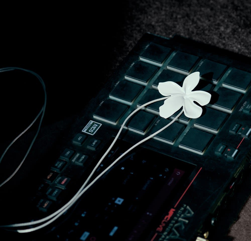 Baltos gėlės formos ausinės guli ant juodo muzikos pado su tekstūruotais mygtukais, susijungdamos su įrenginiu ilgam balto laido juosta.