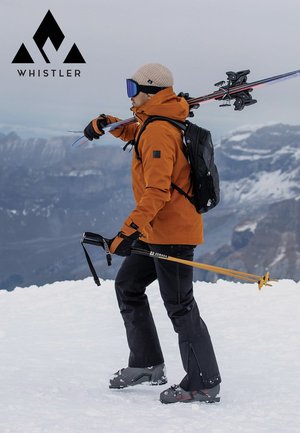 Orangefarbene wasserdichte Jacke, schwarzer Rucksack, schwarze Hose, Schneestiefel, hält Skier und einen Eispickel, steht im Schnee vor bergiger Kulisse.