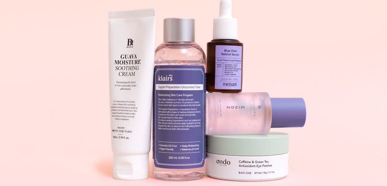 Cinco productos para el cuidado de la piel sobre un fondo rosa: un tubo blanco, una botella transparente con un líquido rosa, una botella oscura con un cuentagotas y un tarro verde.