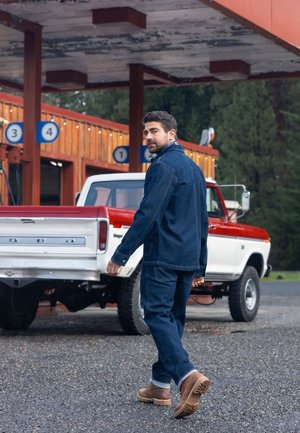 Jeans und eine Denim-Jacke in dunklem Indigo, kombiniert mit braunen Stiefeln, die in der Nähe eines vintage roten und weißen Ford-Trucks an einer Tankstelle spazieren gehen.