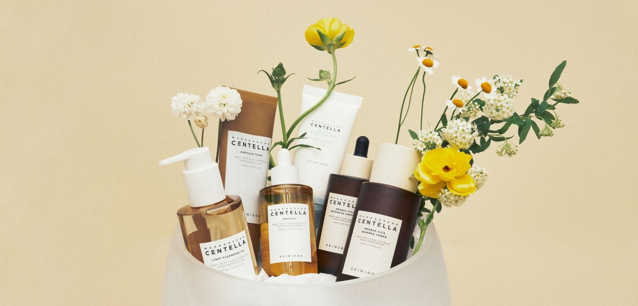 Collezione di prodotti per la cura della pelle in vari flaconi e tubi, con imballaggi marroni, bianchi e trasparenti, accompagnati da fiori gialli decorativi.