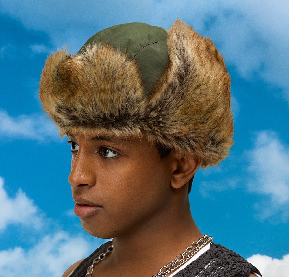 Groene stoffen hoed met een brede, harige rand in een lichtbruine tint. Heeft een ronde bovenkant en een strakke pasvorm. De achtergrond toont een blauwe lucht met wolken.