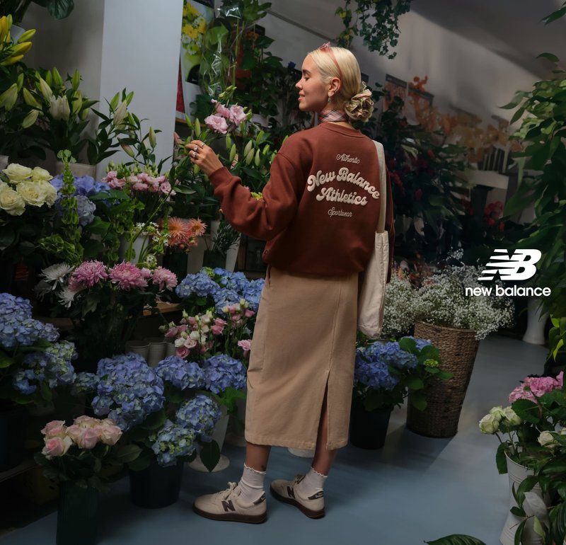 Felpa marrone con scritta bianca, gonna color crema e sneakers beige. Circondata da fiori colorati in un negozio di fiori. Borsa a tracolla semplice.