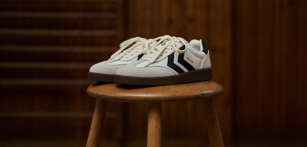 Sneakers with a cream suede upper, black leather accents, white laces, and a brown rubber sole, displayed on a wooden stool.