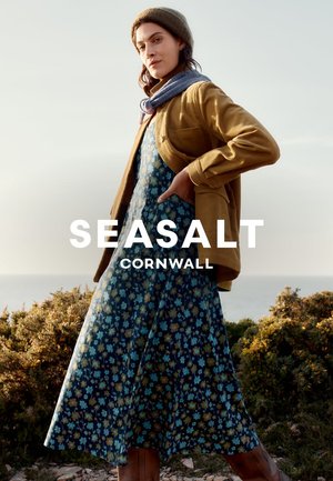 Robe florale avec des motifs bleus et jaunes, superposée sous une veste en corduroy marron. Le modèle porte une écharpe grise et des bottes marron dans un cadre naturel.
