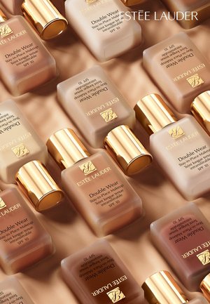 Butelki podkładu Estee Lauder Double Wear w różnych odcieniach skóry, z metalowymi złotymi nakrętkami i etykietami. Gładka szklana tekstura, ułożone na beżowym tle.
