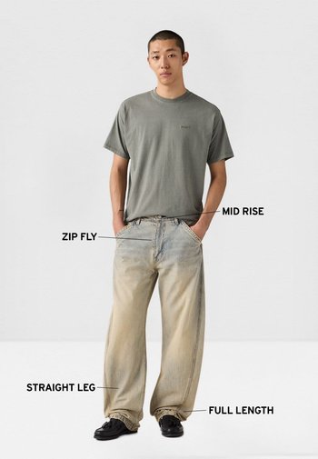 Hombre con camiseta gris y vaqueros de lavado claro, de corte recto, tiro medio, cremallera y largo completo, de pie con las manos en los bolsillos.