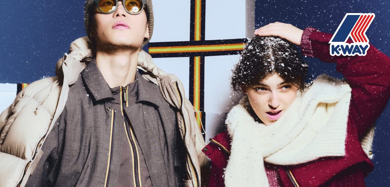 Dos modelos llevan ropa de abrigo de invierno: uno con una chaqueta acolchada beige, el otro con un abrigo rojo y una amplia bufanda blanca. A su alrededor cae nieve.