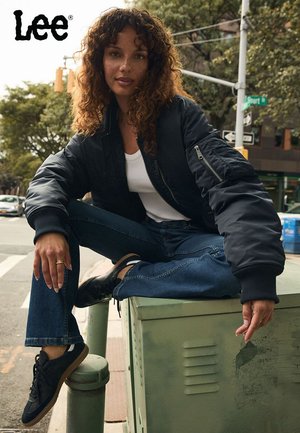 Svart bomberjakke med glidelåsdetaljer, hvit tanktop, blå jeans og svarte sneakers med gummisåler, sitter på en grønn verktøykasse.