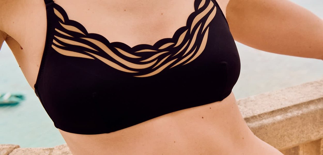 Persona apoyada en una barandilla de piedra con un top de bikini negro con un diseño intrincado de tirantes retorcidos a lo largo del escote.