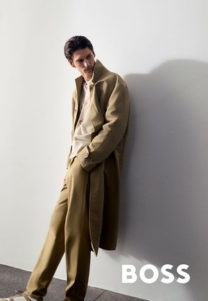 Man in beige trenchcoat en bijpassende broek leunt tegen een witte muur, kijkt naar beneden, met het "BOSS"-logo rechtsonder.