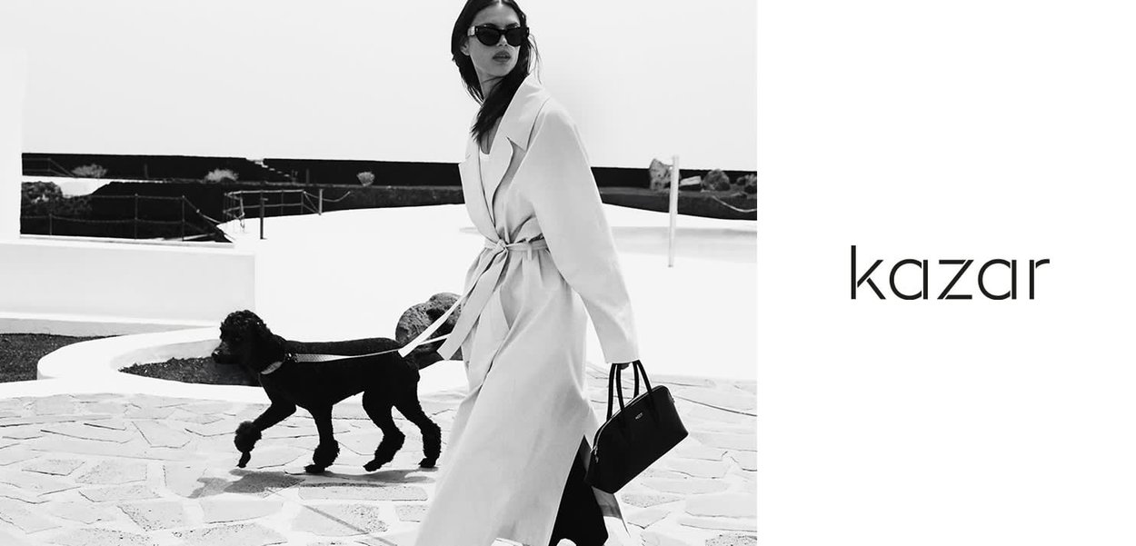 Light-colored trench coat with a belt, paired with sunglasses and a black handbag, walking a black dog on a stone path. Minimalist background.