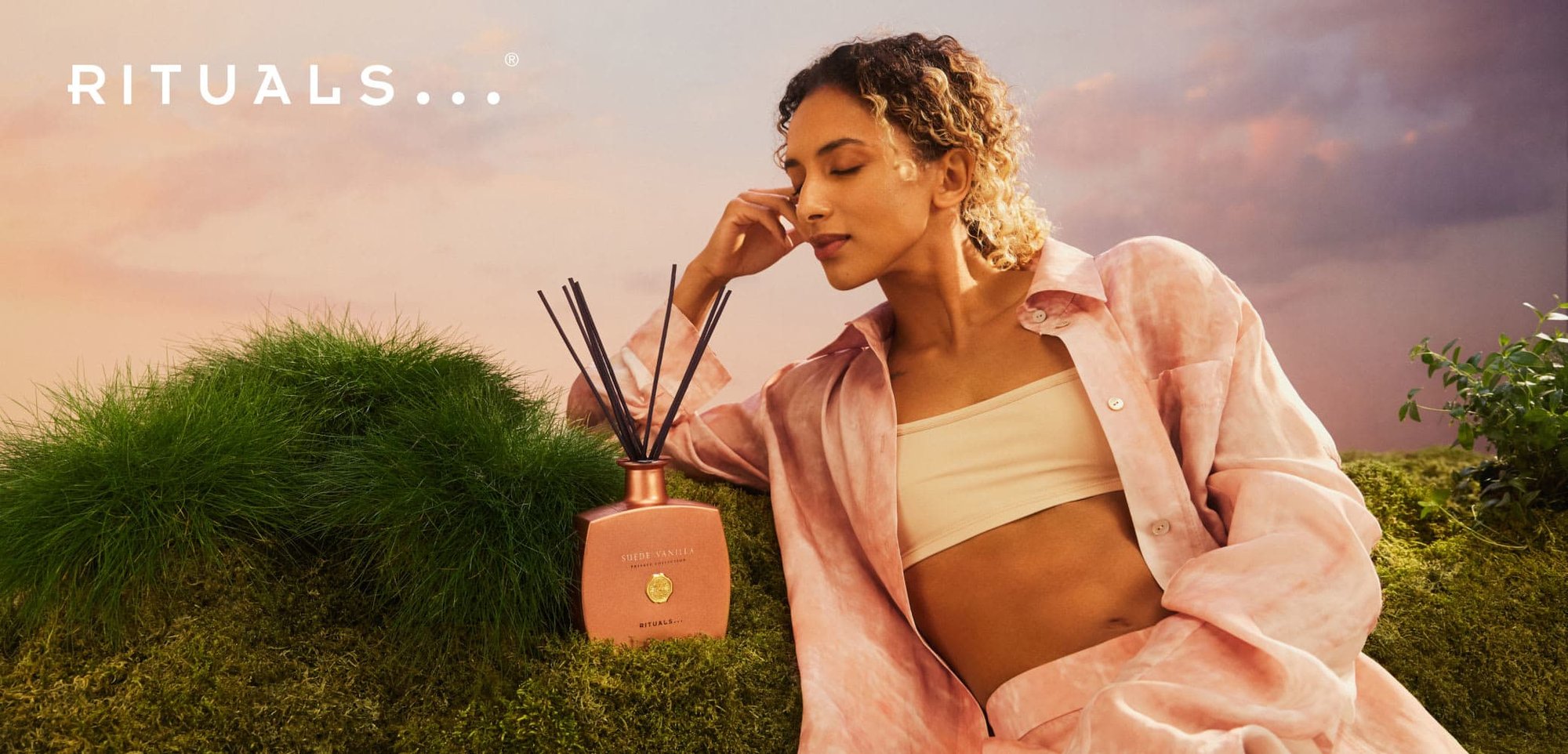 Diffusore Rituals Suede Vanilla con bottiglia in rosa oro opaco e canne nere, posizionato su un prato verde e uno sfondo caldo e morbido.