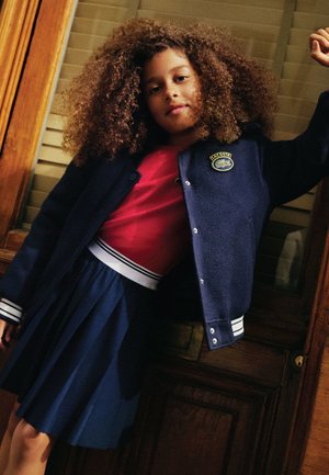 Giacca in pile blu navy con logo, camicia rossa e gonna plissettata blu navy. Presenta dettagli a strisce bianche sulle maniche e sull'orlo della gonna.