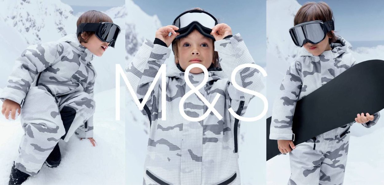 Enfant en combinaison de ski camouflage blanc-gris et lunettes de ski noires pose dans un décor montagneux enneigé, tenant un snowboard dans une main.