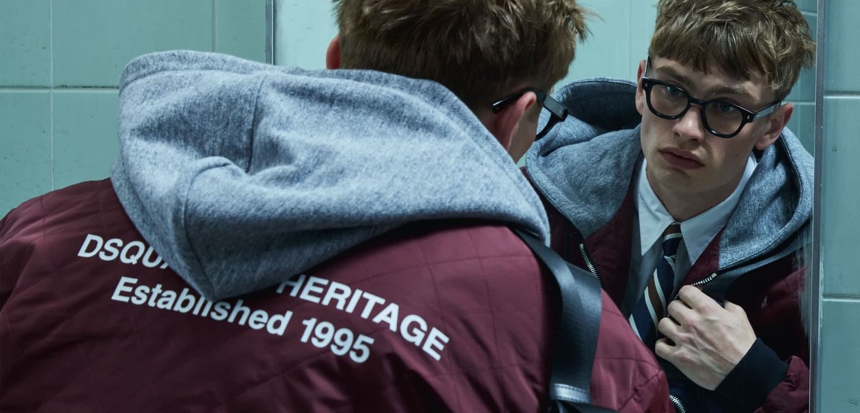 Joven con gafas ajusta una chaqueta burdeos con capucha gris frente a un espejo, texto "HERITAGE Establecido en 1995" visible en la parte trasera.