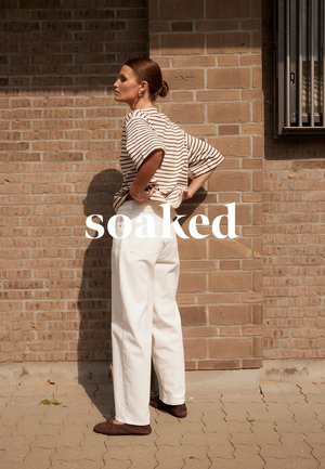 Femme en pantalon blanc et chemise rayée, les mains sur les hanches, debout contre un mur de briques, avec le texte « trempée » centré au-dessus d'elle.