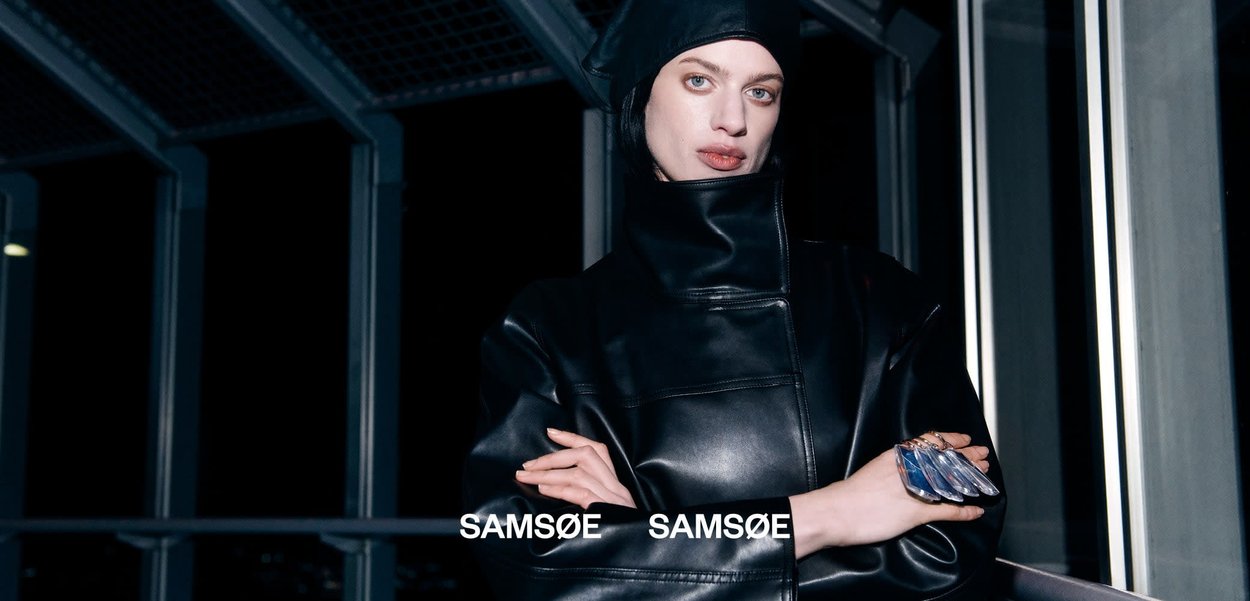 Kvinne i svart skinnjakke med hette står med armene krysset i et moderne, mørkt metall- og glassarkitektonisk miljø om natten.