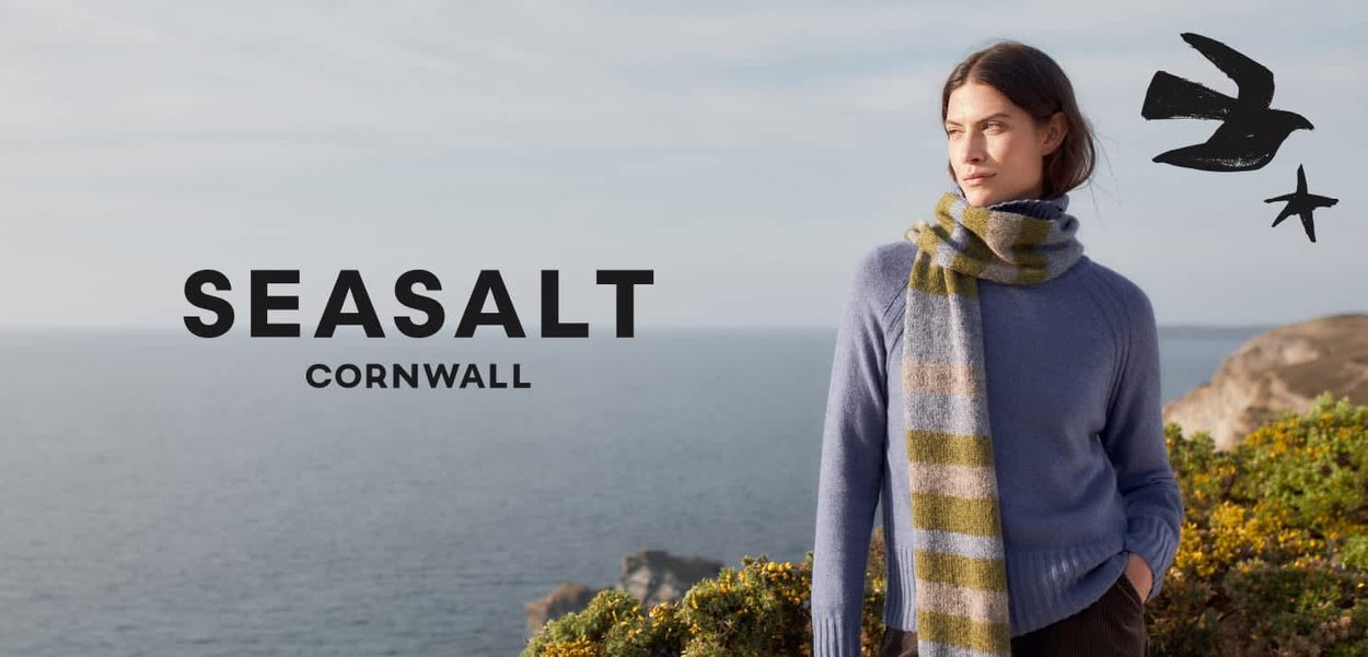 Femme en pull bleu et écharpe rayée debout sur une falaise surplombant la mer avec le texte "SEASALT CORNWALL" et des illustrations d'oiseaux à proximité.