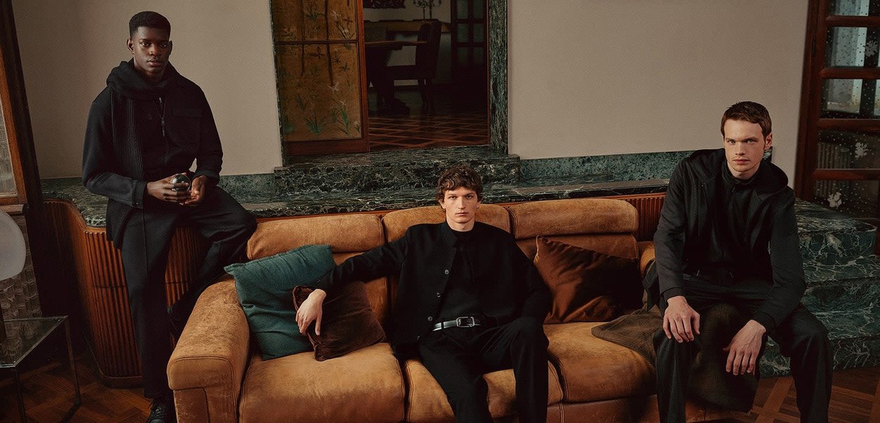 Drei Models in dunkler Kleidung sitzen auf einem braunen Ledersofa mit grünen Akzenten. Der Hintergrund zeigt strukturierten Wände und Holzelemente.