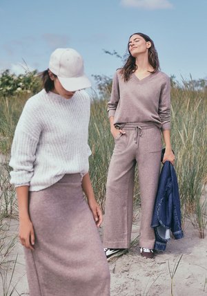 Lichtgrijs gebreid sweater, mauve wijde broek en een bijpassende rok. Eén model houdt een spijkerjack vast, de andere draagt een pet. Zandachtige achtergrond.