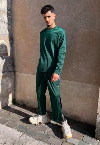 Grön, långärmad topp och matchande byxor med tryckknappar. Löst sittande, slät textur. Klumpiga vita sneakers med ljusbruna sulor.