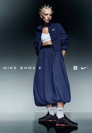 Nike sportkleding met een marineblauwe trackjack, witte crop top en een marineblauwe ballonrok. Zwarte Nike Shox Z-schoenen en grijze sokken maken de look compleet.