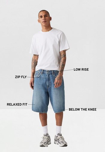 Shorts de mezclilla azul de tiro bajo con un ajuste relajado, cierre de cremallera y dobladillo por debajo de la rodilla. Combinados con una camiseta blanca lisa y zapatillas.