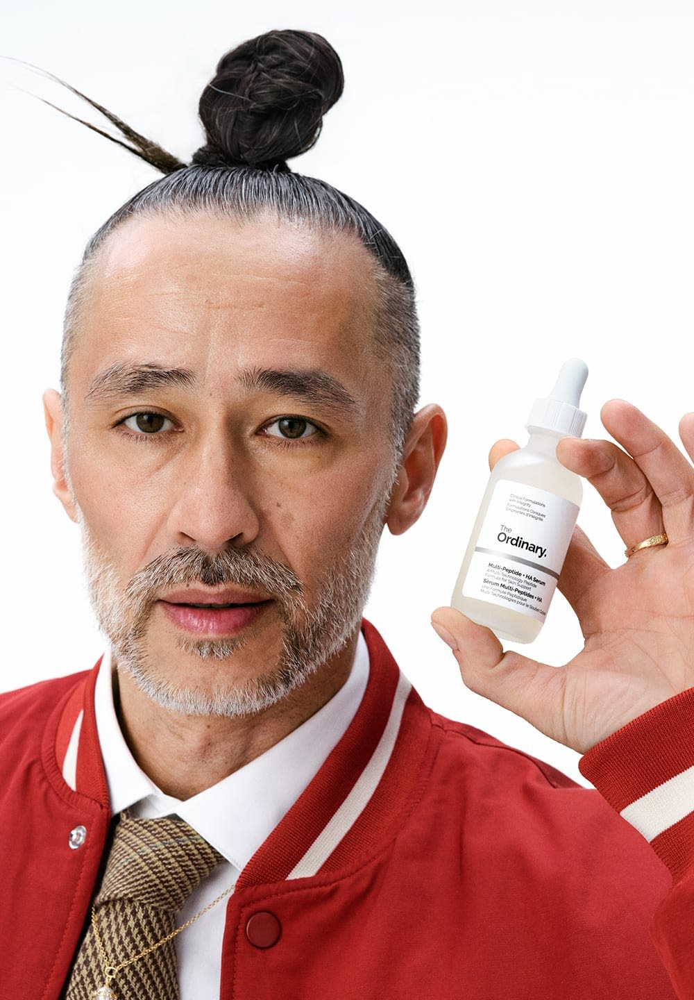 Clear serum bottle with dropper, white label, black text. Model in red jacket with white trim holds bottle, hair styled in a bun.