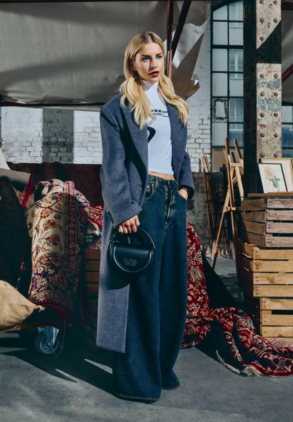 Cappotto grigio oversize, top attillato bianco, pantaloni in denim blu larghi e borsa tonda nera. Lo sfondo presenta motivi e texture.
