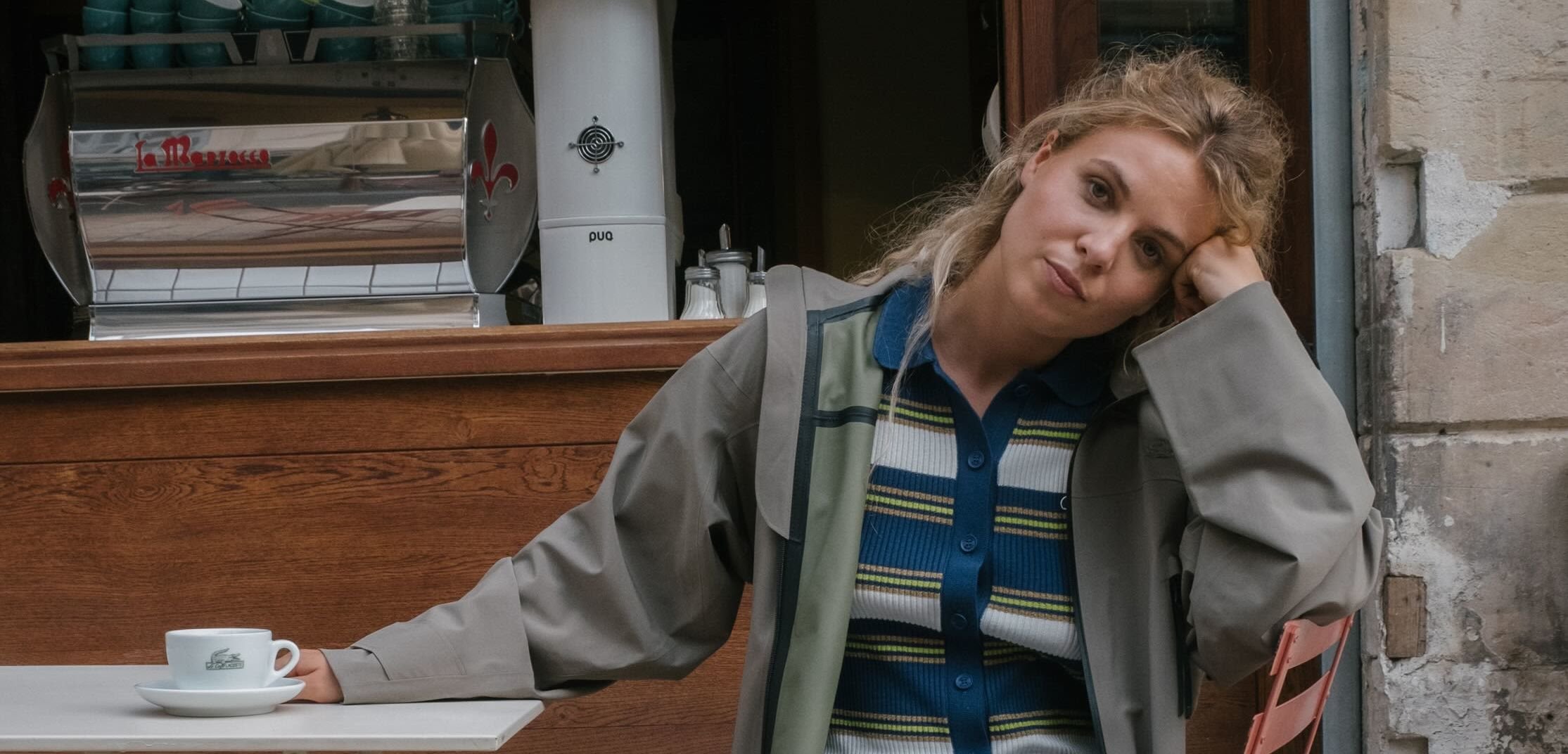 Eine weiße keramische Kaffeetasse auf einer Untertasse steht auf einem Tisch. Im Hintergrund sind eine silberne Espressomaschine und eine Holztheke zu sehen.