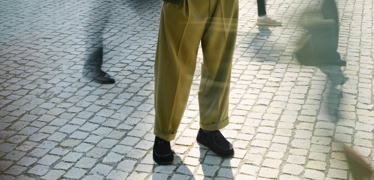 Schwarze Schuhe, hellbraune weite Hosen und eine strukturierte Oberfläche, mit verschwommenen Figuren, die auf einer gepflasterten Straße gehen.