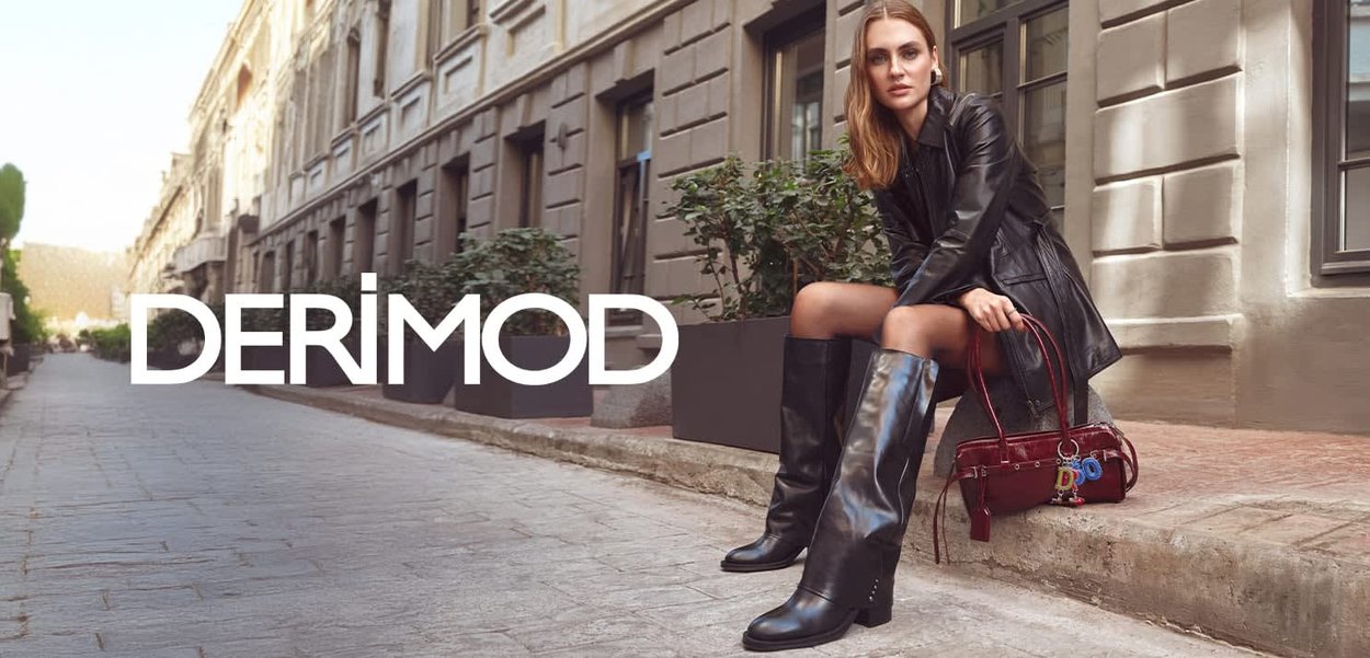 Femme en manteau en cuir noir et bottes montant jusqu'aux genoux assise sur un trottoir en pierre tenant un sac à main bordeaux dans une rue de la ville avec des bâtiments et des plantes.