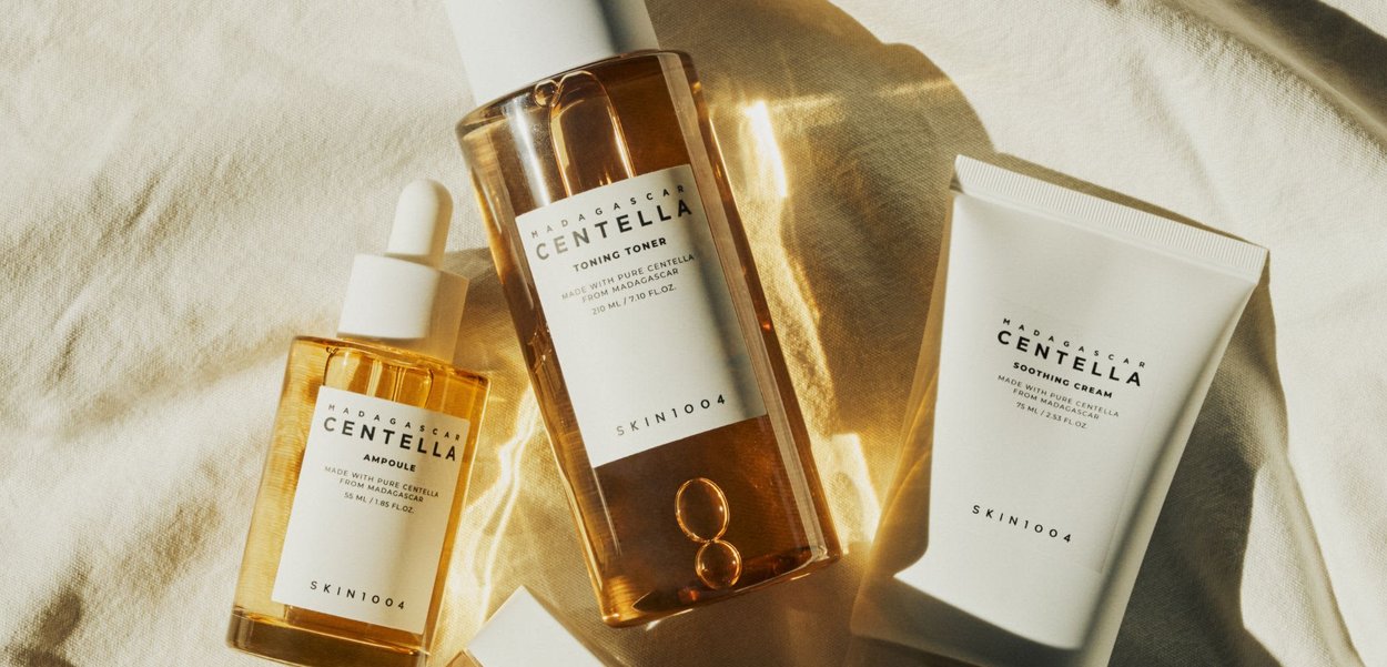 Three skincare products on a light fabric: a transparent bottle of toning toner, a yellow ampoule, and a white soothing cream tube.
