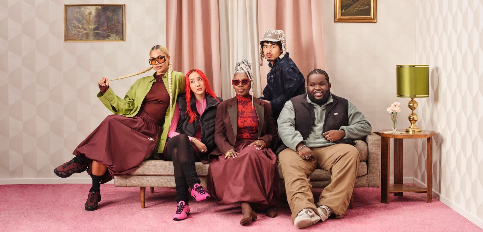 Fünf Personen sitzen auf einem Sofa in einem Raum mit pinkem Teppich und gemusterten Tapeten. Sie tragen unterschiedliche Outfits, darunter eine grüne Jacke, rotes Haar und Sonnenbrillen.